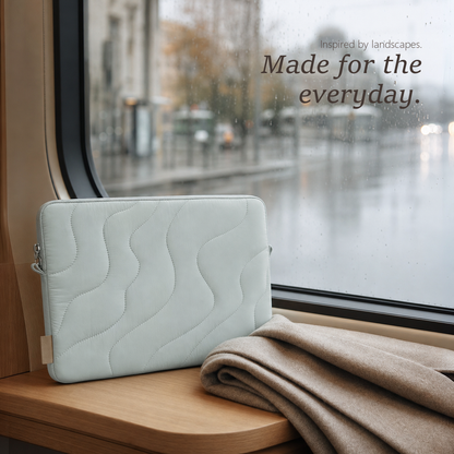 TerraSleeve laptop sleeve in cloud mist, resting on a train window ledge with a folded beige blanket against a rainy city view.